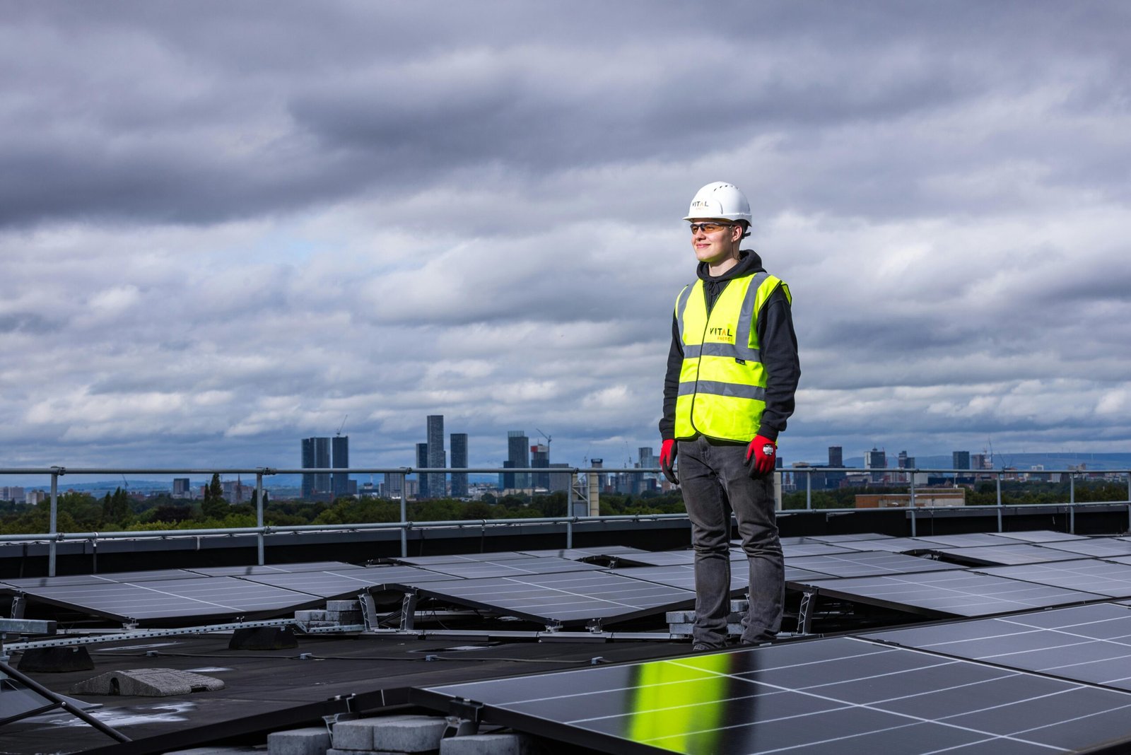 Engineer standing on rooftop inspecting solar panels with city skyline in background.