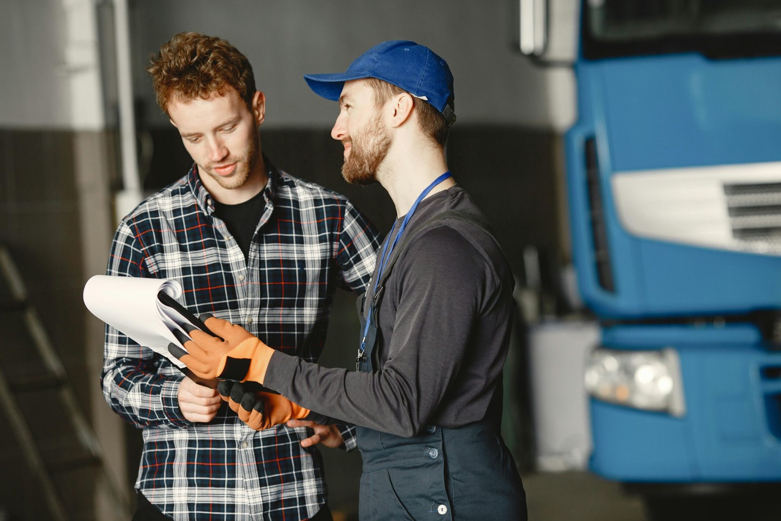 Mechanic in uniform consults with customer in garage setting beside a blue truck.